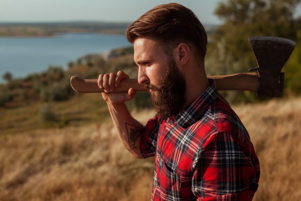 A man in a plaid shirt holding an axe over his shoulder with a view of a river behind him.