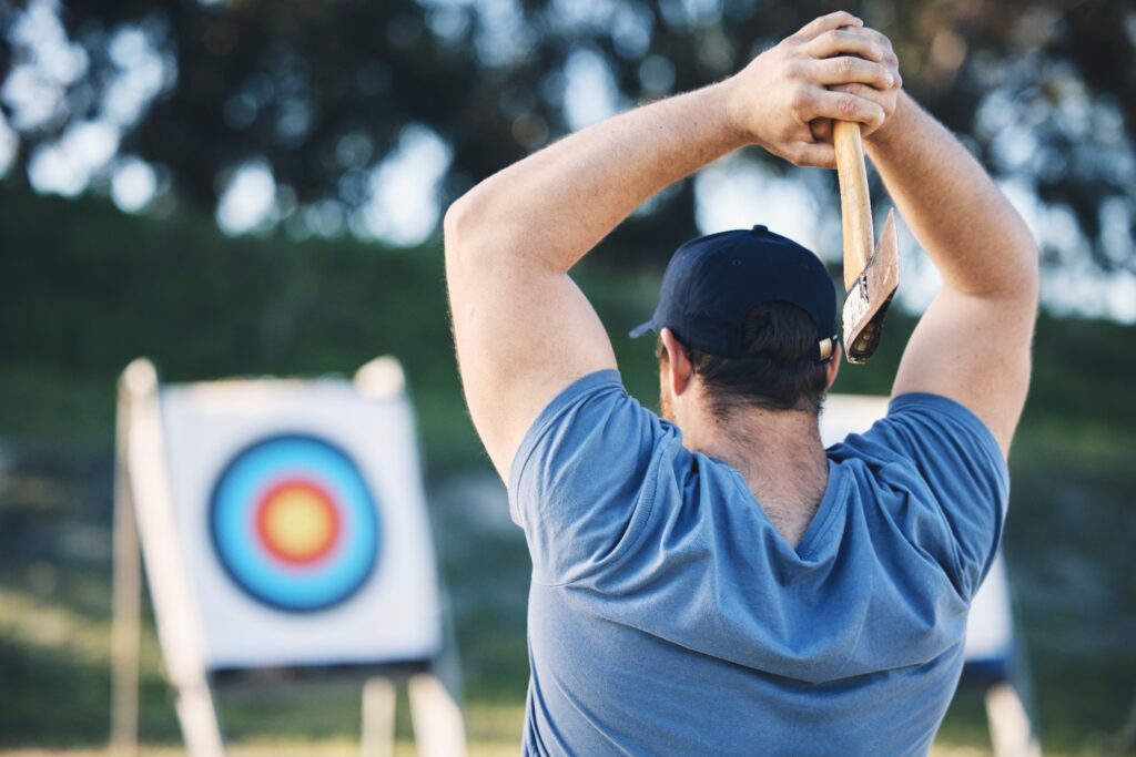 A man holds an axe over his head, preparing to throw it at a target in an outdoor sports range.