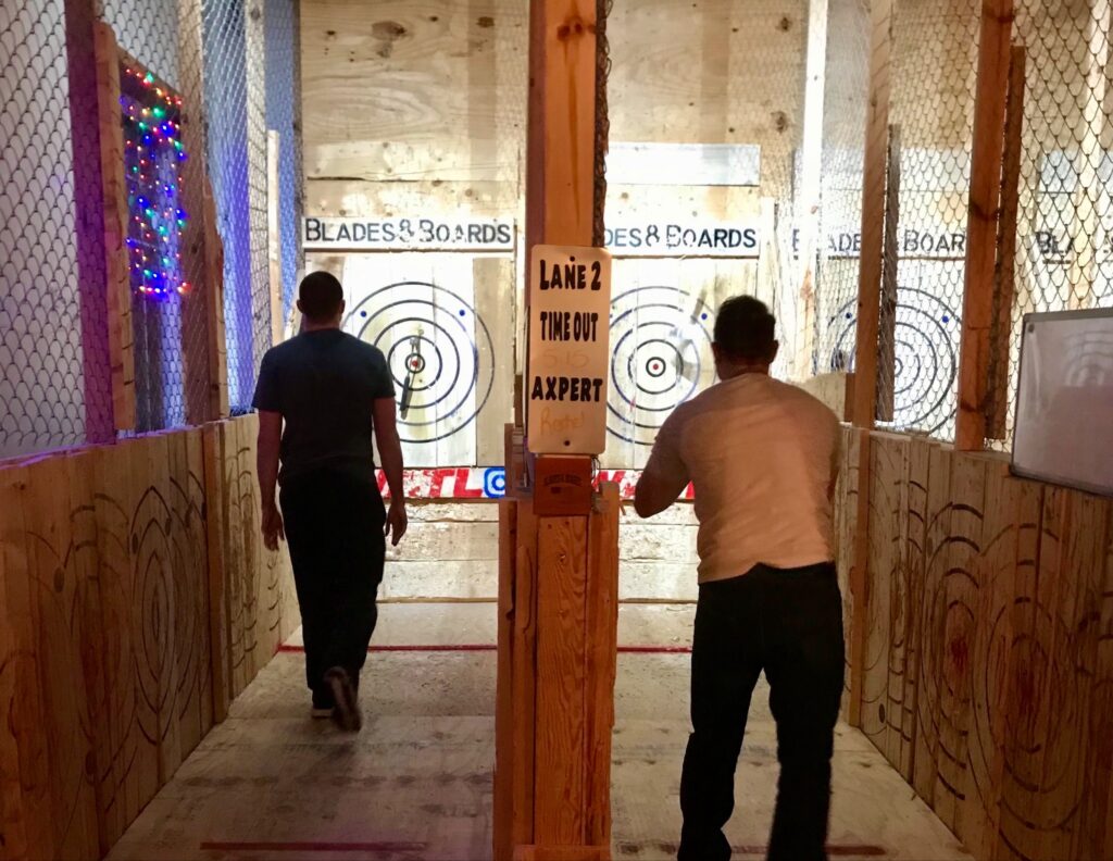 Two men throw axes side-by-side in an indoor range.