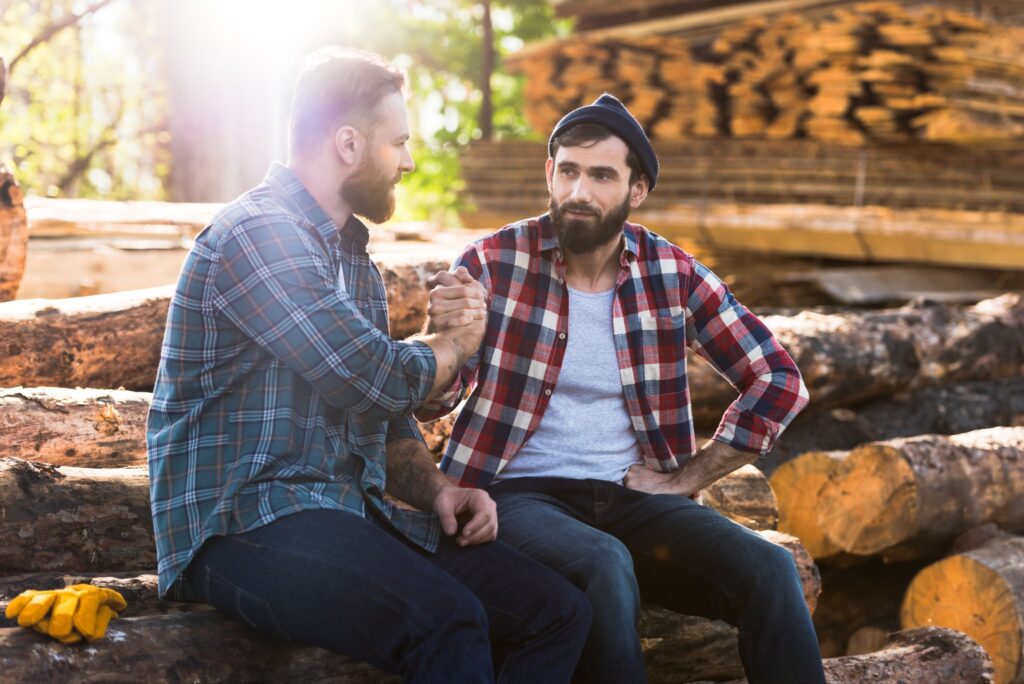 Two lumberjacks sit on a stack of logs together and shake hands.