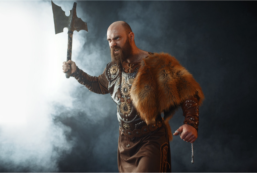 A bearded viking man wearing a belt holds up a viking axe.
